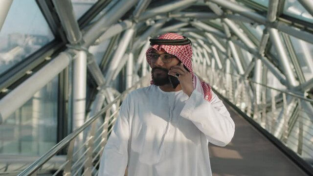 Medium slowmo of young wealthy Arab sheikh talking on smartphone while walking along indoor glass bridge of high-class office building with beautiful city view