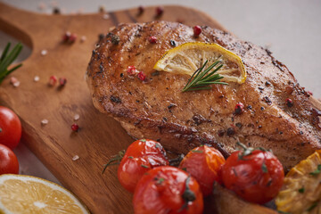 Grilled chicken breast on a wooden board and vegetables with tomatoes and mushrooms, potato, garlic, herbs, lemon, rosemary, salt  on a stone background with space for text