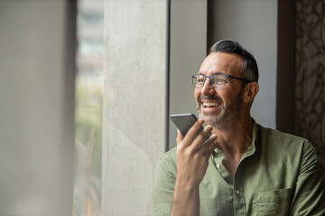 Smiling mature man with beard talking on phone next to sunny window