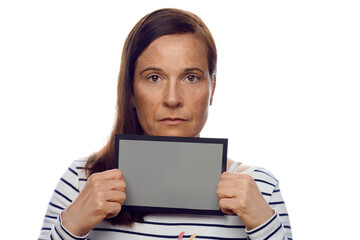 Middle-aged woman holding up a blank grey card with border to her chin while looking at the camera with a serious thoughtful expression isolated on white
