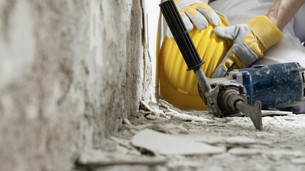 house renovation concept, construction worker holds in hands helmet, close up with jackhammer and wall plaster rubble on background