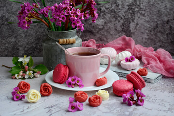 Appetizing macarons, a cup of tea, pink flowers and chocolate in the form of roses on a gray textured background. Spring still life