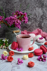 Vertical spring composition. A cup of tea, appetizing macarons, pink flowers and chocolate in the form of roses on a gray textured background