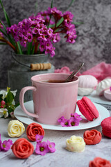 Fototapeta na wymiar Close-up of a pink cup of tea and appetizing macarons, beautiful flowers on a gray textured background. Vertical