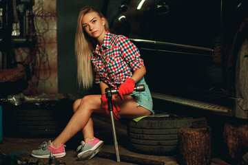 A girl in a bright plaid shirt and short denim shorts sits on a car wheel with a key to unscrew the wheels at a service station. Handsome mechanic at work.