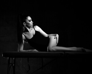 Black and white of young sexy brunette woman in black bodysuit lying on massage table, touching her smooth silky skin on hip after cosmetic procedure, anti-cellulite therapy over dark background