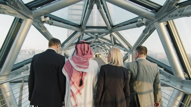 Rear-view medium tilt-up slowmo of team of diverse male and female business partners walking along long empty indoor glass walkway of modern high-class office building