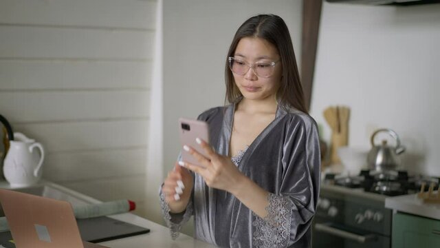Confident slim beautiful Asian woman putting on eyeglasses and taking selfie on smartphone in home office. Portrait of positive young millennial in pajamas enjoying remote working in kitchen indoors