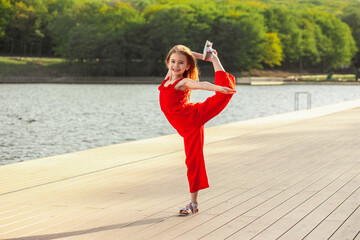 Happy girl in a red jumpsuit on the waterfront making workout. Enjoyment of nature, freedom, life after quarantine.