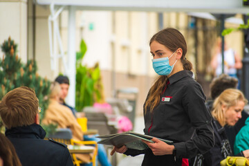 Pretty nice waitress with mask and menu serving clients at the table of an outdoor bar, cafe or...