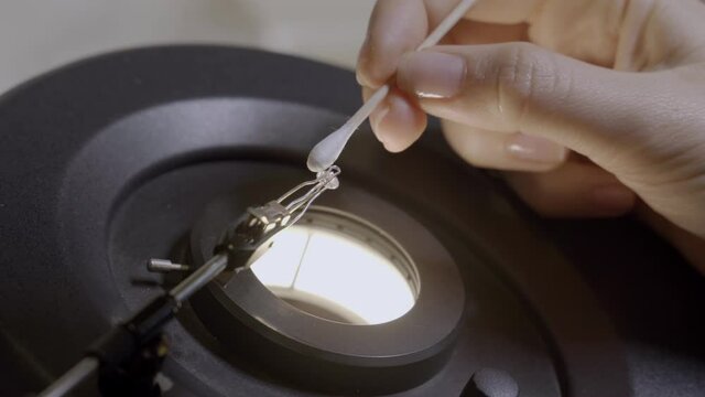Female hands cleaning a diamond under a microscope using cotton-tipped swab. Round shape diamond in professional gemology laboratory under light being meticulously cleaned with a soft q tip