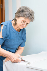 Senior woman dressmaker cutting cloth indoors. Smiling seamstress with glasses at work at home. Hobby or retired work. Vertical photo