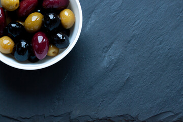 Different kinds of organic ripe olives on oil in the white bowl on the dark stone background. Copy space