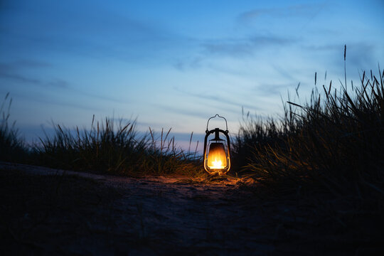 Old classic oil lantern burning with an orange flame on the beach. Sunset sky on background.