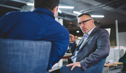 Fototapeta na wymiar Caucaisan male boss in optical eyeglasses and formal suit explaining information to colleague discussing brainstorming process at desktop, businessmen in spectacles have conversation in office