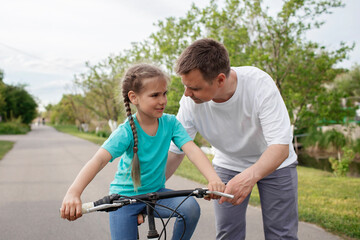Obraz premium Father teaching her little daughter to ride a bicycle, happy summer in village, healthy lifestyle, childhood, fatherhood and upbringing, concept for father day