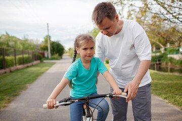Obraz premium Father teaching her little daughter to ride a bicycle, happy summer in village, healthy lifestyle, childhood, fatherhood and upbringing, concept for father day