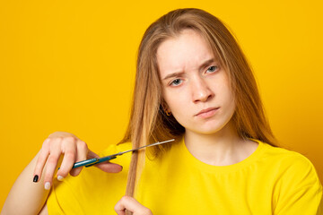 Sad and unhappy teen girl cutting her hair with scissors while standing on yellow background. Young student experiments with her hairstyle at home.