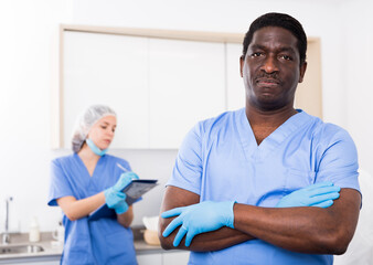Fototapeta premium Portrait of confident african american male cosmetologist standing in modern aesthetic medicine office on background with female assistant filling out medical form..