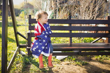 American girl Toddler child wrapped herself in the USA flag on Independence Day. A child runs with...