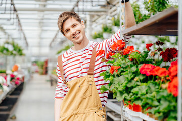 Young agro-engineer posing near cart with geranium flowers in small pots for sale..