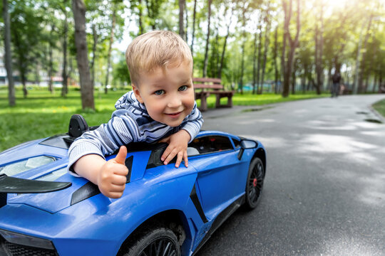 Portrait of cute little caucasain blond toddler boy enjoy having fun riding electric powered toy car by asphalt path road city park at summer day. Happy child show thumbs up gesture playing outdoors