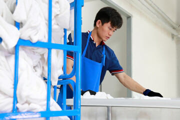 A laundry staff wearing a dark blue polo shirt is checking towels. Blur trolley cart with towels inside is on the foreground. Selective focus on model's face. Shot taken in the factory.