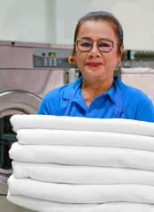 Woman wearing a blue laundry uniform is holding a stack of clean towels and smiling at camera. Shot taken in the factory.
