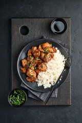 Spicy teriyaki chicken fillet pieces with rice, green onions and black sesame seeds on black plate on a dark slate, stone or concrete background. Top view with copy space.