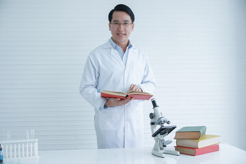 Asian Scientist man with white coat and glasses holding and reading text book with microscope for...