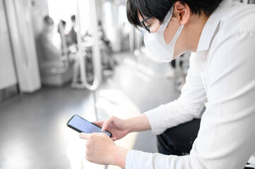 Fototapeta premium Asian man wearing surgical face mask using smartphone on skytrain or urban train. Coronavirus (COVID-19) infection prevention in public transportation. Health awareness for pandemic protection