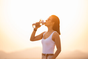 woman taking a break to drink from water bottle while hiking and poles standing on a rocky mountain ridge looking out valleys and peaks in a healthy outdoors lifestyle concept. explorer young woman.