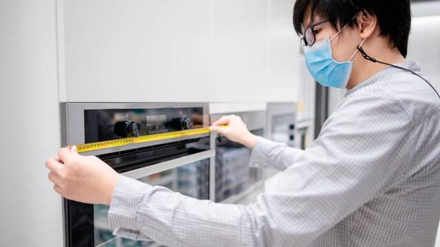 Asian Man Interior Designer Wearing Face Mask Using Tape Measure On Oven In Kitchen Showroom At Furniture Store. Handy Cooking Appliance For Domestic Kitchen. Home Improvement Concept