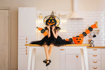 Funny blonde girl kid8-9 years old in a witch costume sitting on the kitchen table decorated for the Halloween holiday. Traditions, holidays, scenery concept.