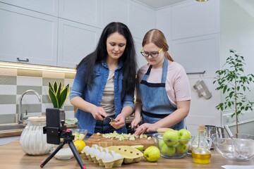 Mom and teen daughter cooking apple pie together, looking at smartphone screen