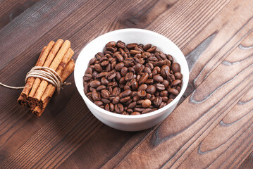Grains of coffee in white bowl and bunch of cinnamon sticks on wooden background