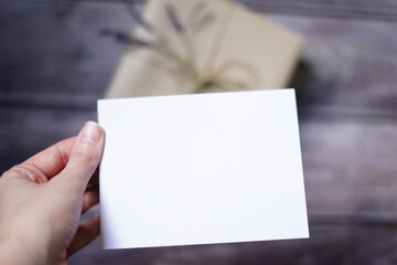 Woman's hand holding blank white card with a present on wooden background. Surprise gift mock-up scene. Empty space for text. Copy space.