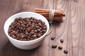 Grains of coffee in white bowl and bunch of cinnamon sticks on wooden background