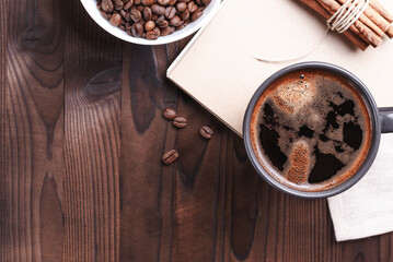 Grains of coffee in white bowl, cup of black coffee and bunch of cinnamon sticks on wooden background