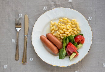 Boiled sausages with pasta and vegetable salad on a plate on the table. Top view.