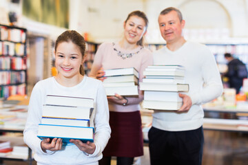 Fototapeta na wymiar Positive smiling preteen girl and her parents holding piles of books bought in bookstore