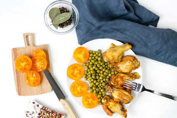 Chicken legs baked in the oven with green peas and yellow tomatoes on a white plate with a fork. Top view with a copy of the space