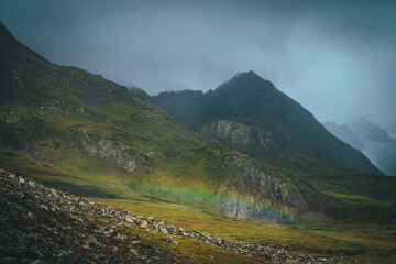 Dramatic rainy alpine landscape with low rainbow in green valley and dark sharp pinnacle in low clouds. Pointed rocks in overcast weather. Atmospheric awesome view to pointy mountain in low clouds.