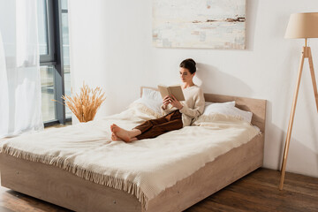 barefoot young woman reading book while chilling on bed at home