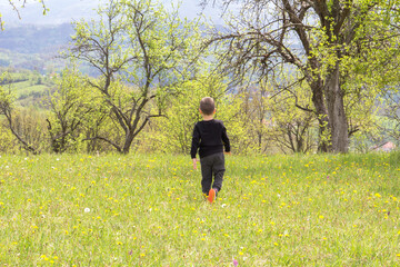 A small child with his back turned is playing in the meadow. He enjoys the beautiful nature and the view of the distant hills.