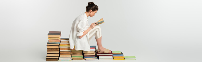side view of young woman in sandals reading while sitting on pile of books on white, banner