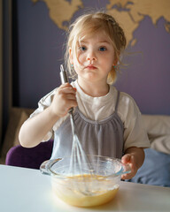 Portrait of cute blue eyed girl looking at camera while cooking omelet, learning how to prepare food while spending leisure time at home, cute small child whipping eggs in bowl with whisk for pie