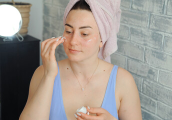 Beautiful young woman applies moisturizer to her face. Woman doing beauty treatments at home. Home facial care concept. 