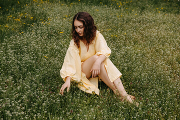 Young barefoot woman wearing a yellow summer dress, sitting in a field with green grass.