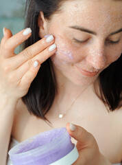 Beautiful young woman applies violet scrub to her face. Woman doing beauty treatments at home. 